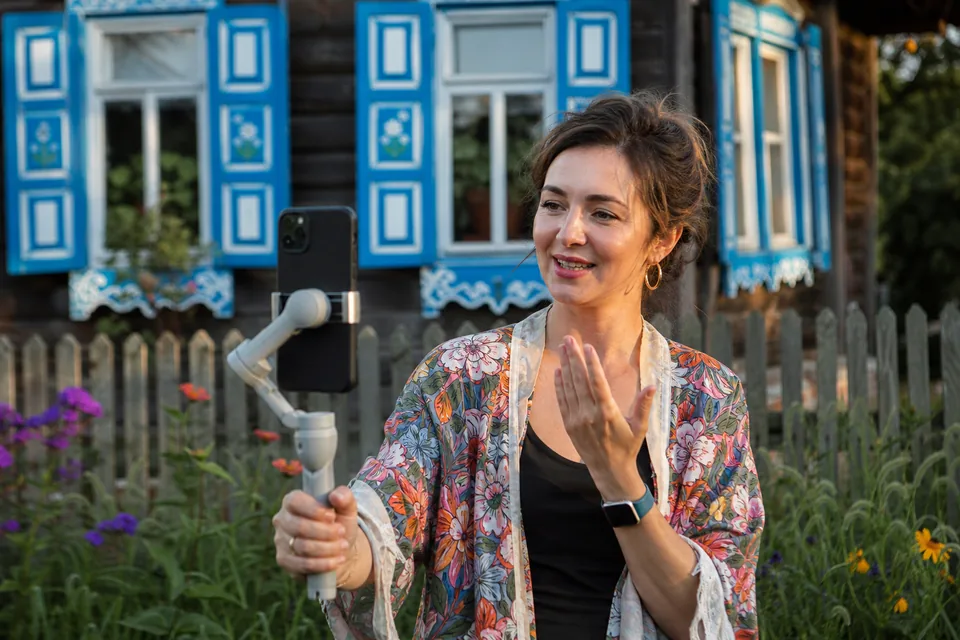 Twórczyni nagrywa telefonem film przed drewnianym domem z malowanymi okiennicami na Podlasiu